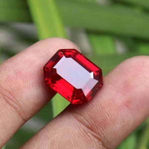 Puede incluir: Una gema de color rojo intenso y vibrante tallada en forma de esmeralda rectangular con bordes facetados. La piedra muestra una gran claridad y una superficie pulida, reflejando la luz a través de sus múltiples facetas angulares.