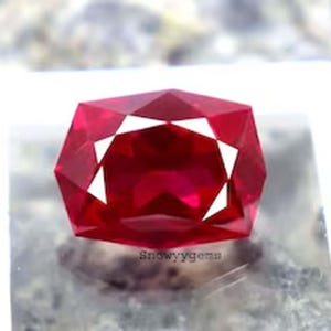 May include: A vibrant, cushion-cut red gemstone with a faceted surface that reflects light. The stone displays a deep, saturated crimson hue and is positioned above a small, light-colored base featuring the text Snowyygems in black lettering.