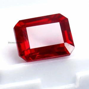 Puede incluir: Una gema roja de corte rectangular con superficies facetadas que reflejan la luz, mostrando tonos carmesí profundos y claridad interna. El texto 'Snowyygems' está impreso en pequeñas letras negras en el lado izquierdo de la piedra.