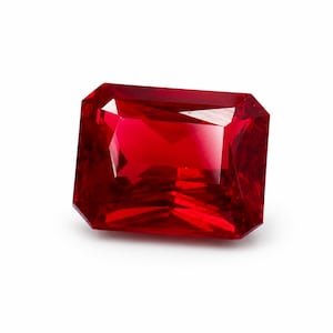 Puede incluir: Una gema de color rojo intenso y vibrante, tallada en forma rectangular con superficies facetadas que reflejan la luz. La piedra presenta bordes afilados y limpios con un acabado transparente y pulido, mostrando un rico tono carmesí en toda su estructura.