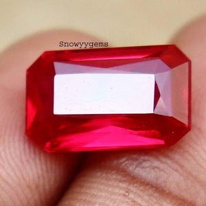 May include: A rectangular, faceted gemstone with a vibrant, saturated red color. The stone features a clear, polished surface with sharp edges and geometric facets that reflect light. The text "Snowyygems" is printed in black above the gemstone.