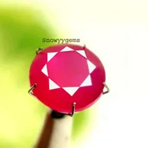 May include: A round, vibrant pink gemstone featuring a star-shaped facet pattern on its surface. The stone is secured by four metal prongs and displays the text Snowyygems above it. The gem has a smooth, polished finish and a bright, saturated color.