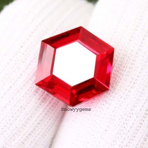 May include: A vibrant red gemstone cut into a precise hexagonal shape with multiple facets that reflect light. The gemstone features a clear, polished surface and is positioned against a white textured fabric. The text Snowyygems is printed below.
