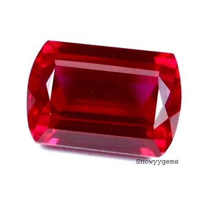 May include: A vibrant, rectangular-cut gemstone with a deep, saturated red color and faceted surfaces that reflect light. The stone features sharp, clean edges and a polished finish. The text 'Snowyygems' is printed in small black letters at the bottom.