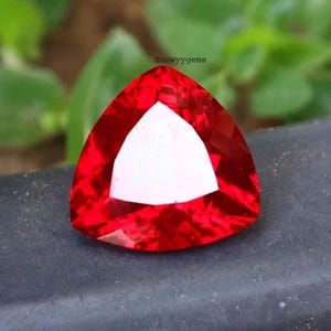 May include: A vibrant red gemstone cut into a triangular shape with multiple facets that reflect light. The stone features a brilliant, saturated crimson hue and a smooth, polished surface. The text Snowyygems is visible above the gemstone.