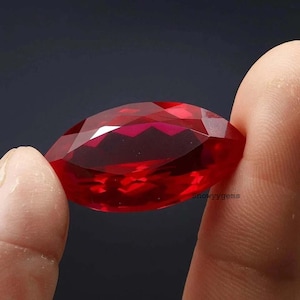 May include: A faceted, oval-shaped, transparent red gemstone with a brilliant cut. The stone displays deep crimson hues and sharp geometric facets that reflect light. The text 'snowyygems' is printed in small black letters on the surface of the gem.
