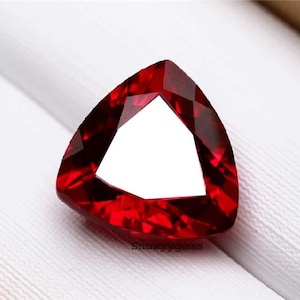 May include: A vibrant red gemstone cut into a triangular shape with multiple facets that reflect light. The stone features a deep crimson hue and a polished surface. The text Snowyygems is printed in small black letters at the bottom center.