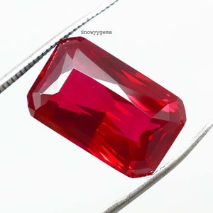 May include: A vibrant, rectangular-cut red gemstone held by silver-toned metal tweezers. The faceted stone displays deep crimson hues and internal light reflections. The text "Snowyygems" is printed in small black letters above the gemstone.