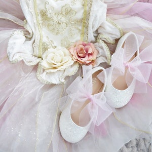 Può includere: Un tutù di tulle bianco con bordi dorati e un corpetto bianco con ricami dorati. Due scarpe bianche scintillanti con fiocchi rosa sono sopra il tutù.