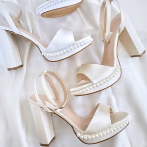May include: Four pairs of white satin platform heels with block heels and ankle straps. Each shoe features a row of small pearls along the platform edge. The shoes are arranged on a white satin surface.