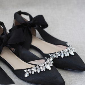 May include: Black satin bridal flats with a pointed toe and a large crystal embellishment on the vamp. The shoes have a black satin ribbon that ties around the ankle.