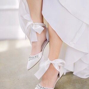 Puede incluir: Bailarinas de sat&eacute;n blanco con punta, adornos de cristal y lazos en el tobillo. Los zapatos tienen un tac&oacute;n bajo y est&aacute;n adornados con delicados lazos de sat&eacute;n blanco. Zapatos de novia o de ocasi&oacute;n especial.