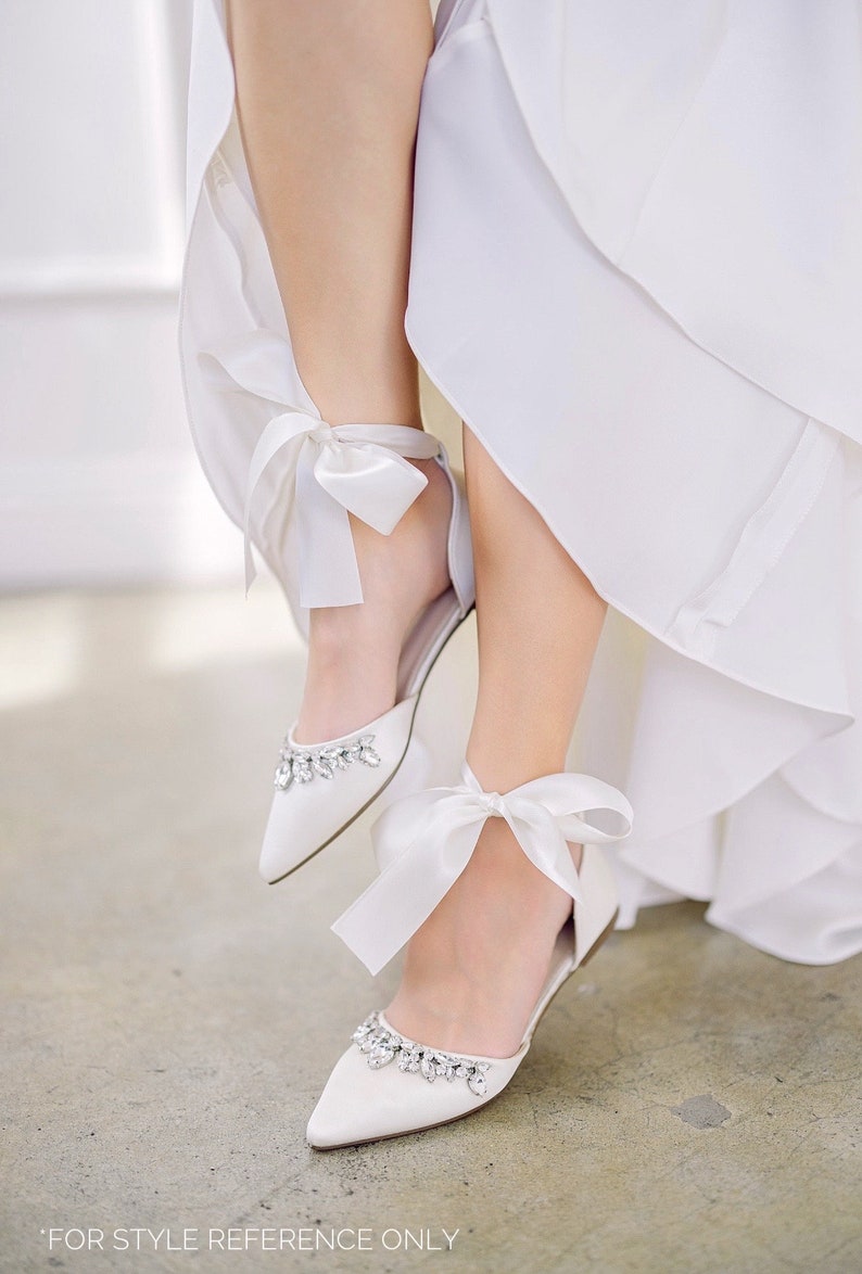Puede incluir: Bailarinas de sat&eacute;n blanco con punta, adornos de cristal y lazos en el tobillo. Los zapatos tienen un tac&oacute;n bajo y est&aacute;n adornados con delicados lazos de sat&eacute;n blanco. Zapatos de novia o de ocasi&oacute;n especial.