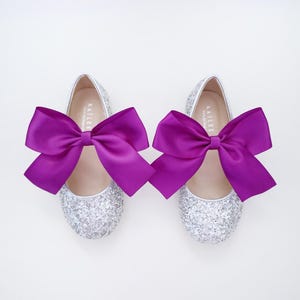 May include: A pair of silver glitter ballet flats with purple satin bows. The shoes have the word "Kailee" written on the inside.