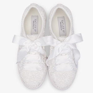 May include: A pair of white lace-covered sneakers with satin ribbon laces and bows. The sneakers have a low profile and a white sole. The brand name "KAILIE P" is printed on the insole.