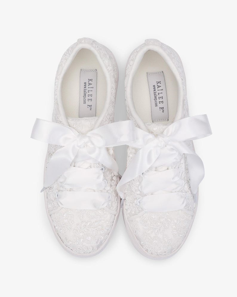 May include: A pair of white lace-covered sneakers with satin ribbon laces and bows. The sneakers have a low profile and a white sole. The brand name "KAILIE P" is printed on the insole.