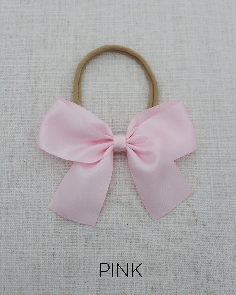 May include: A pink satin bow tied on a nude elastic headband. The headband is for babies and toddlers.