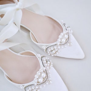 May include: A pair of elegant white satin flats with pointed toes. The shoes are adorned with pearl and rhinestone embellishments around the vamps. Long satin ribbons are tied in bows at the ankles, adding a touch of sophistication.