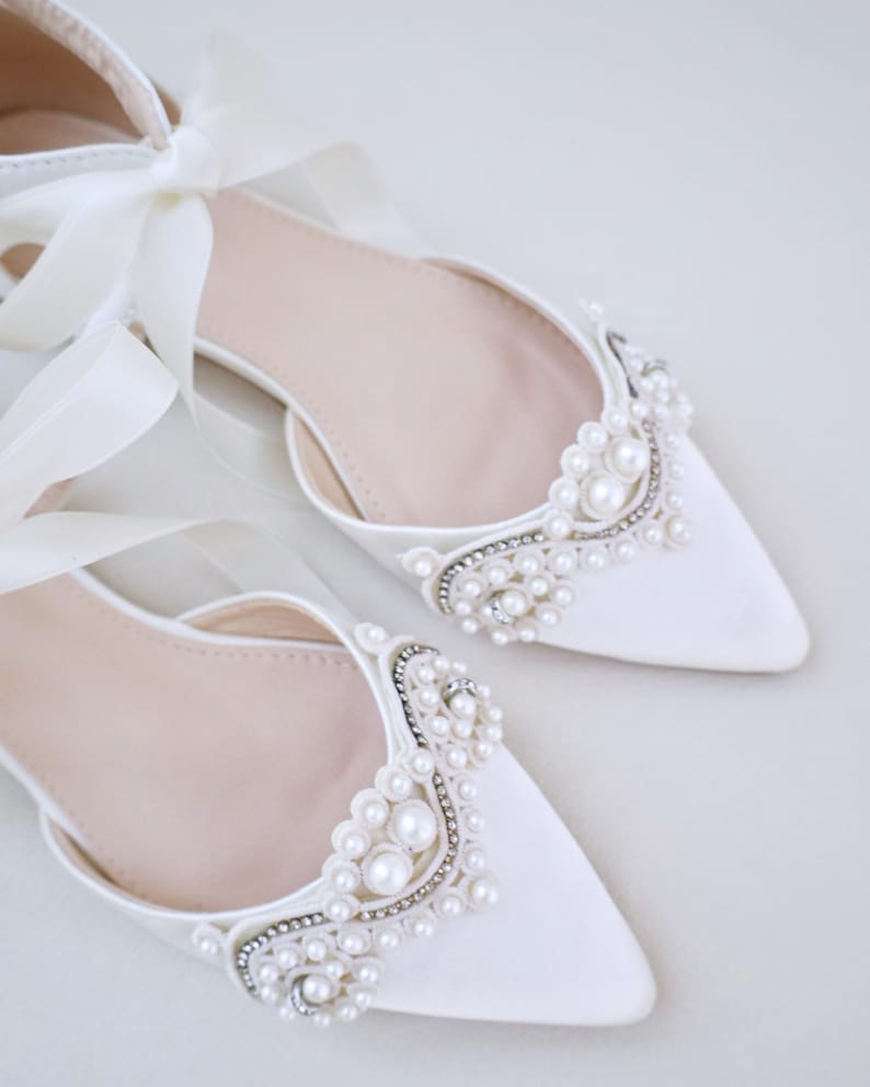 May include: A pair of elegant white satin flats with pointed toes. The shoes are adorned with pearl and rhinestone embellishments around the vamps. Long satin ribbons are tied in bows at the ankles, adding a touch of sophistication.