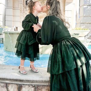 May include: A mother and daughter wearing matching green tulle dresses with a layered skirt. They are kissing in front of a fountain decorated with a Christmas wreath.