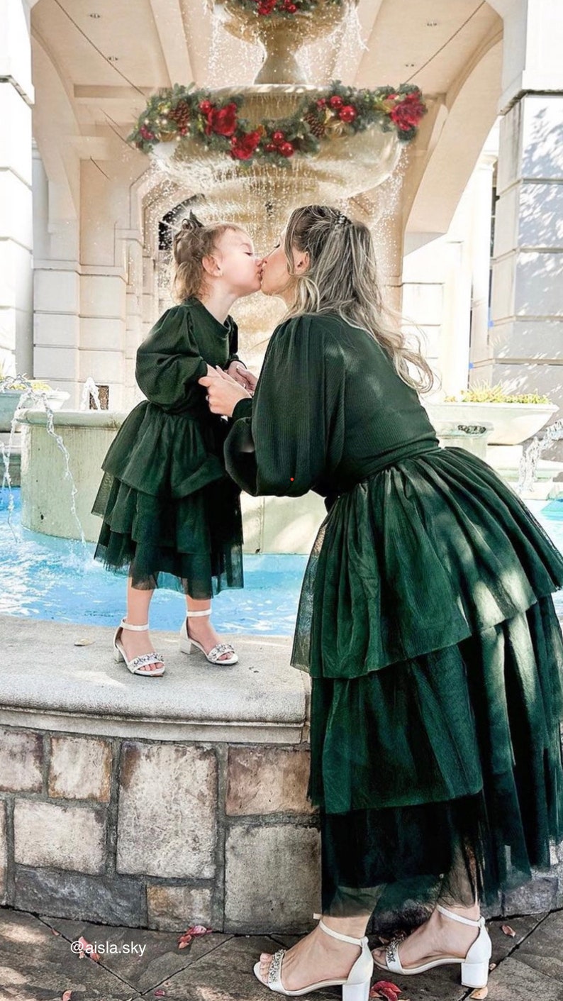 May include: A mother and daughter wearing matching green tulle dresses with a layered skirt. They are kissing in front of a fountain decorated with a Christmas wreath.
