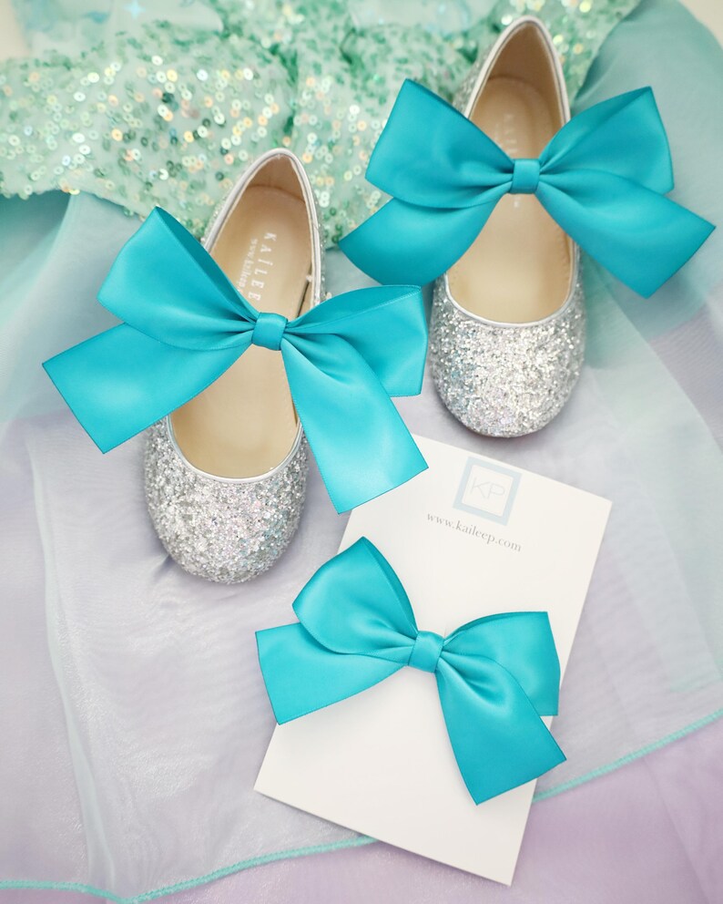 May include: A pair of silver glitter shoes with turquoise satin bows. A matching turquoise satin bow is also included. The shoes have the text "Kailer" on the side. The text "www.kailerp.com" is on a white card.