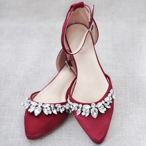 Puede incluir: Un par de bailarinas de sat&eacute;n rojo con punta. Los zapatos tienen una correa en el tobillo y est&aacute;n adornados con pedrer&iacute;a transparente y brillante en el borde superior. Los zapatos est&aacute;n dise&ntilde;ados para ocasiones formales.