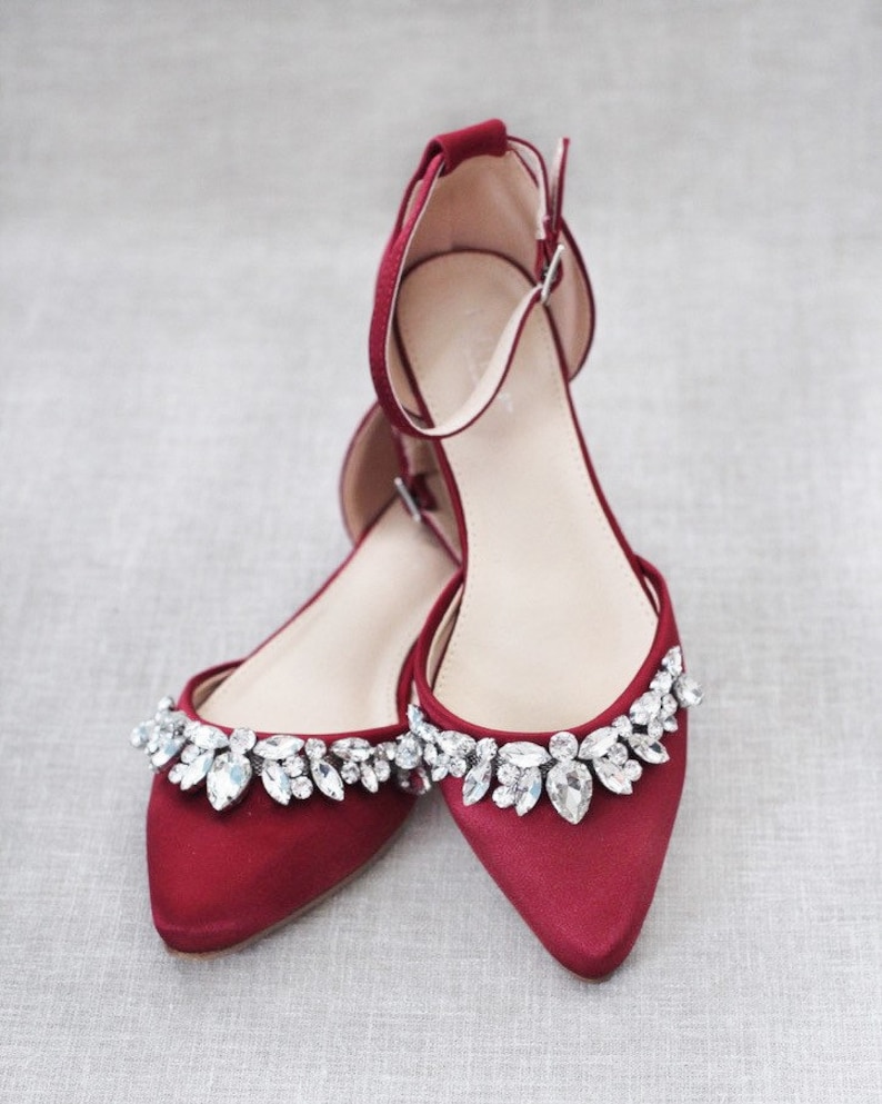 Puede incluir: Un par de bailarinas de sat&eacute;n rojo con punta. Los zapatos tienen una correa en el tobillo y est&aacute;n adornados con pedrer&iacute;a transparente y brillante en el borde superior. Los zapatos est&aacute;n dise&ntilde;ados para ocasiones formales.