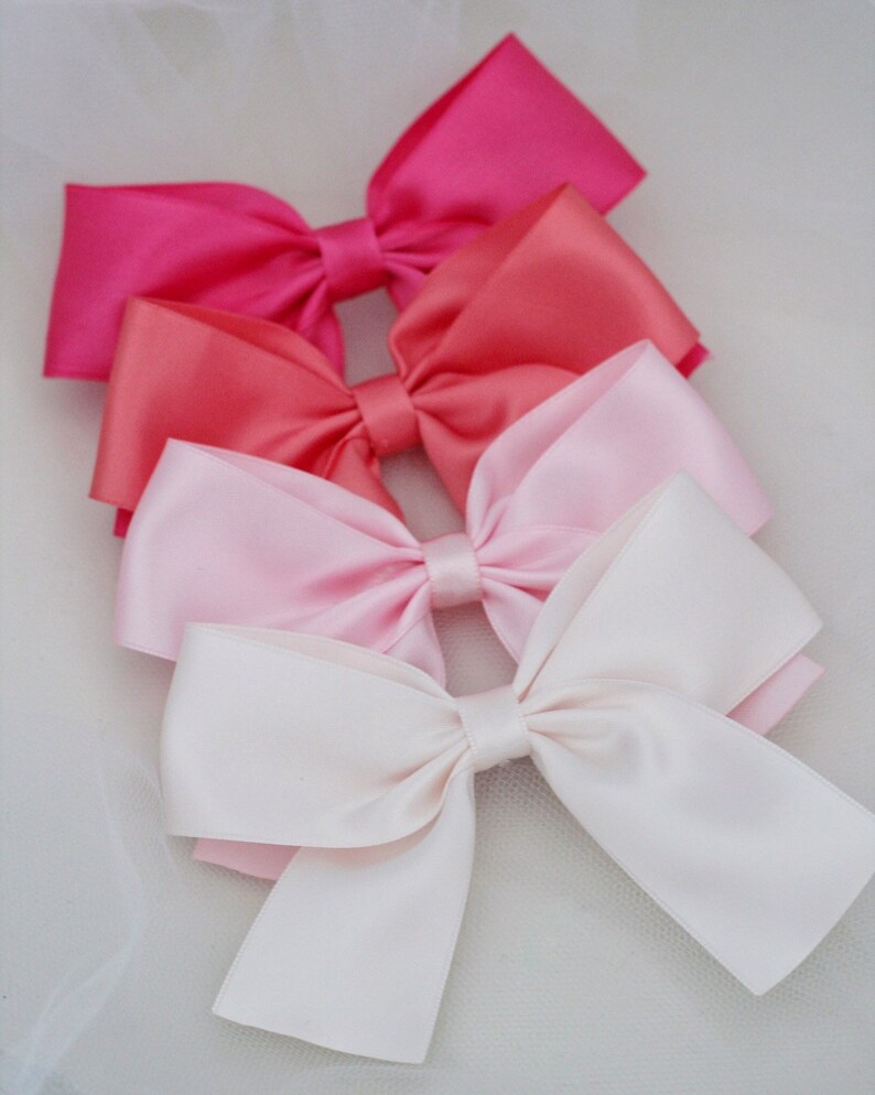 May include: Four satin ribbon bows in shades of pink, coral, and white. The bows are arranged in a stack, with the largest bow at the bottom and the smallest bow at the top.