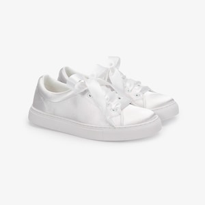 May include: A pair of white satin sneakers with white ribbon laces. The sneakers have a low profile and a white rubber sole. The shoes are set against a white background.