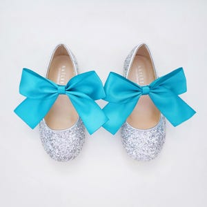 May include: A pair of silver glitter ballet flats with turquoise satin bows. The shoes are tied with a bow at the front.