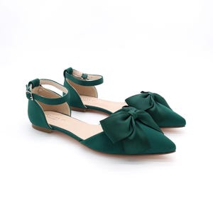 May include: A pair of emerald green satin flats with a large bow on the toe. The shoes have an ankle strap with a buckle closure.
