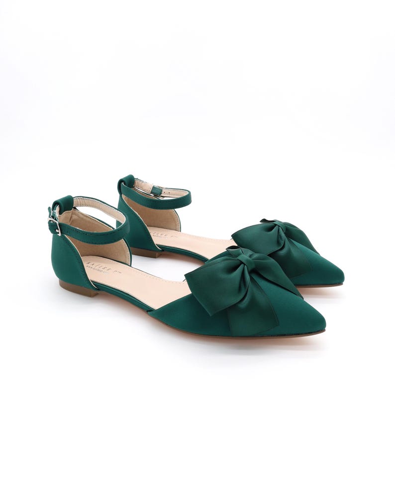 May include: A pair of emerald green satin flats with a large bow on the toe. The shoes have an ankle strap with a buckle closure.