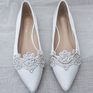 May include: A pair of white satin bridal flats with a pointed toe and a jeweled embellishment on the vamp. The embellishment features a delicate lace design with clear rhinestones.