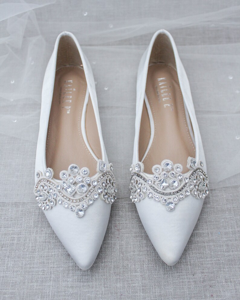 May include: A pair of white satin bridal flats with a pointed toe and a jeweled embellishment on the vamp. The embellishment features a delicate lace design with clear rhinestones.