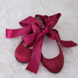 May include: A pair of burgundy satin ballet flats with a large burgundy satin bow tied around the ankles.