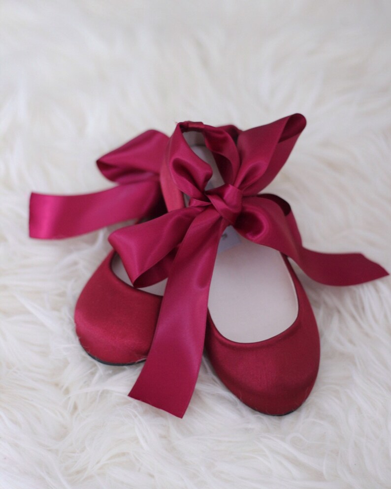 May include: A pair of burgundy satin ballet flats with a large burgundy satin bow tied around the ankles.