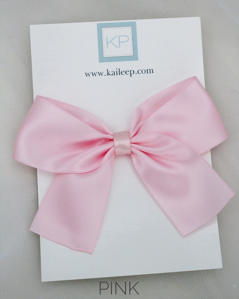 May include: A large pink satin bow with a small centre loop. The bow is on a white card with the text "KP" and "www.kaileep.com" printed on it. The word "PINK" is printed at the bottom of the card.