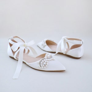 May include: White satin flats with pointed toes and ankle straps. The shoes feature a pearl and rhinestone embellishment on the vamp and long satin ribbons. The flats are designed for a special occasion.