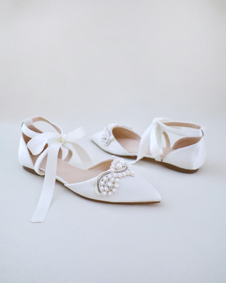 May include: White satin flats with pointed toes and ankle straps. The shoes feature a pearl and rhinestone embellishment on the vamp and long satin ribbons. The flats are designed for a special occasion.