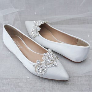 May include: Pair of white satin bridal flats with pointed toes. Each shoe features a large, ornate rhinestone embellishment on the vamp.