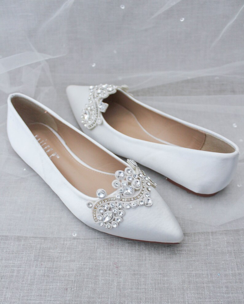May include: Pair of white satin bridal flats with pointed toes. Each shoe features a large, ornate rhinestone embellishment on the vamp.