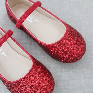 Puede incluir: Un par de bailarinas rojas de purpurina con una tira en la parte superior. Los zapatos tienen un dise&ntilde;o sencillo y son perfectos para una ocasi&oacute;n especial.