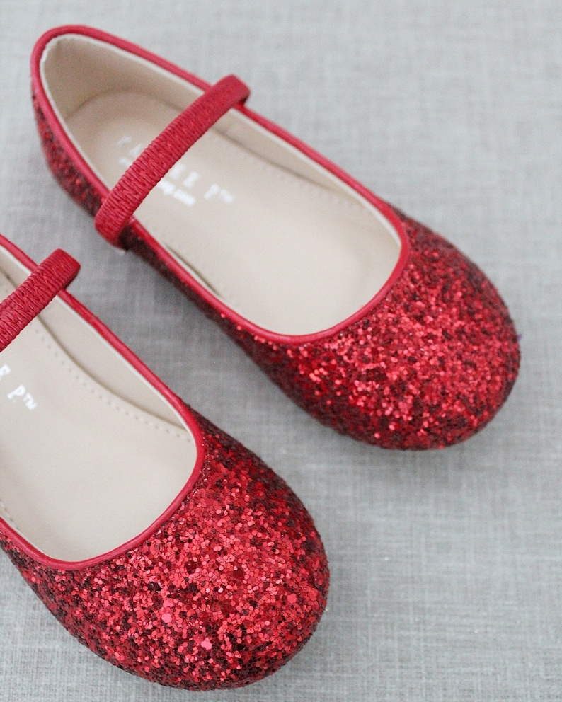 Puede incluir: Un par de bailarinas rojas de purpurina con una tira en la parte superior. Los zapatos tienen un dise&ntilde;o sencillo y son perfectos para una ocasi&oacute;n especial.