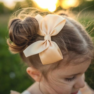 May include: A young girl with brown hair wearing a white dress with a floral pattern. She has a large, cream-coloured satin bow in her hair.