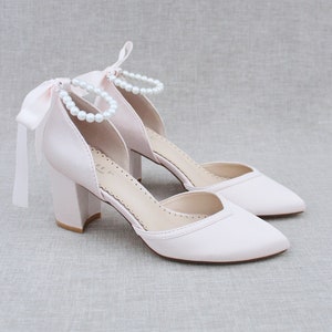 Op de afbeelding: Een paar blush roze satijnen pumps met een blokhak en een parel enkelband. De schoenen hebben een spitse neus en een satijnen strik aan de achterkant.