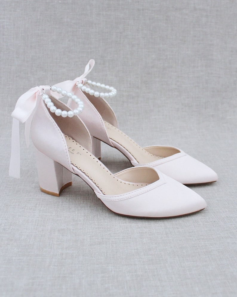 Op de afbeelding: Een paar blush roze satijnen pumps met een blokhak en een parel enkelband. De schoenen hebben een spitse neus en een satijnen strik aan de achterkant.