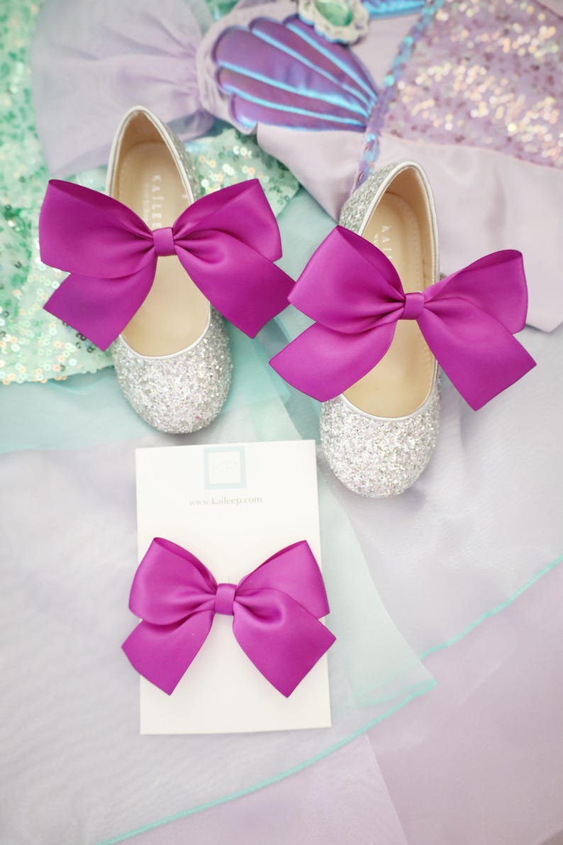 May include: A pair of silver glitter shoes with fuchsia satin bows. The shoes are on a light blue and purple background. A fuchsia satin bow is also shown on a white card with the text "www.kaliebp.com".