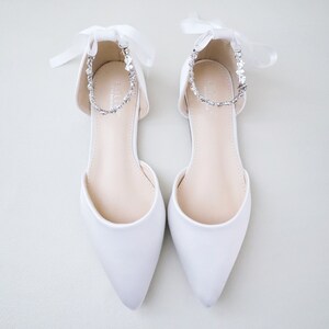 May include: A pair of white satin flats with pointed toes. Each shoe features a delicate ankle strap adorned with sparkling rhinestones and a satin ribbon tie. The shoes are designed for a formal occasion.
