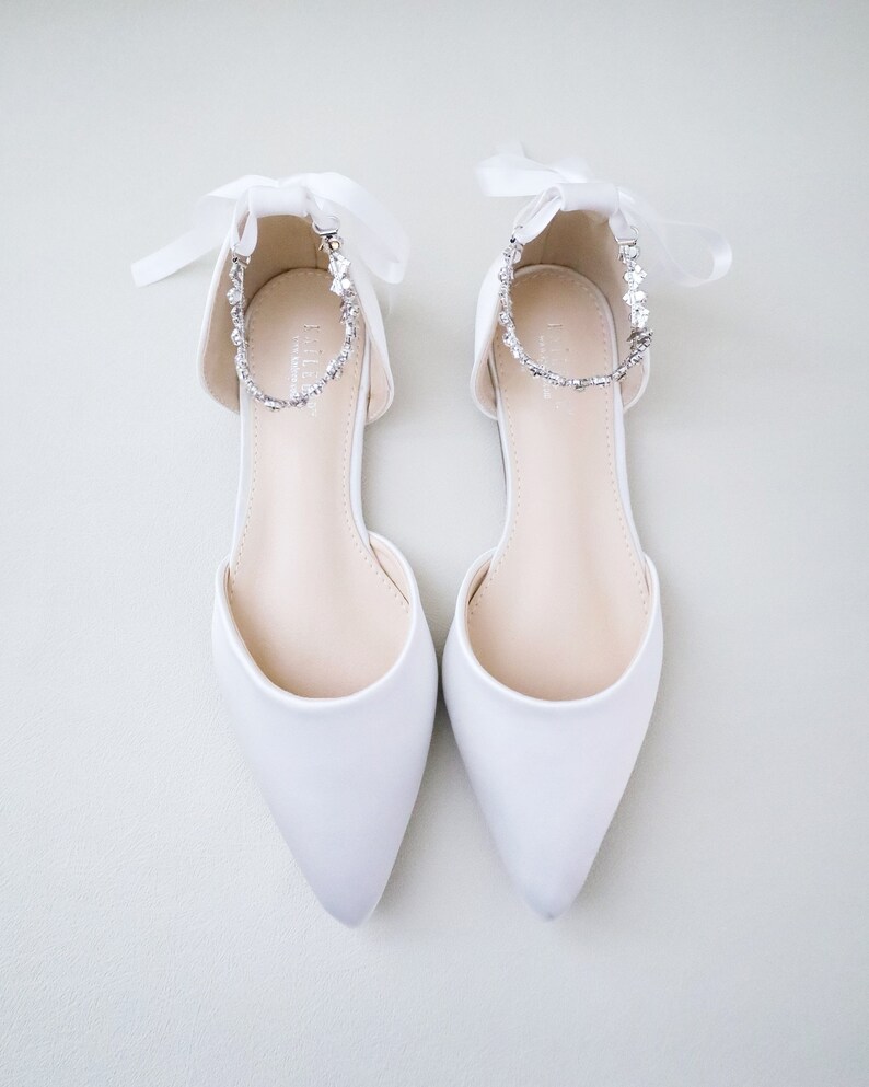 May include: A pair of white satin flats with pointed toes. Each shoe features a delicate ankle strap adorned with sparkling rhinestones and a satin ribbon tie. The shoes are designed for a formal occasion.